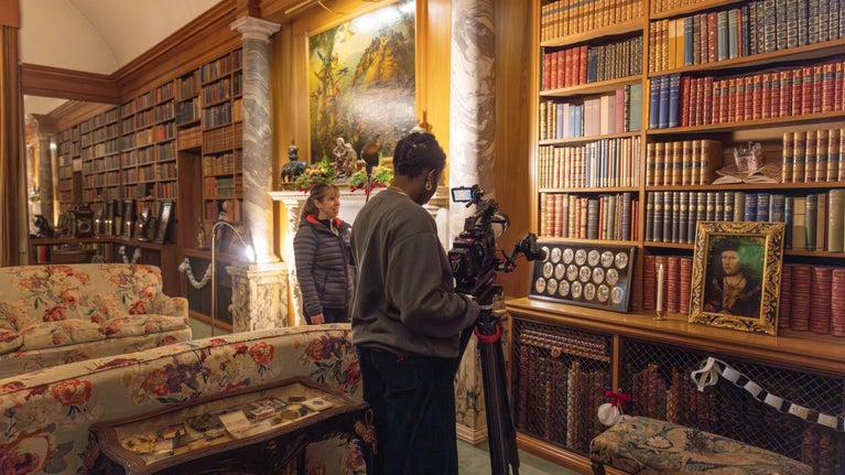 A camera lady is filming decorations in front of the bookshelves in the Anglesey Abbey library. A member of staff is assisting..